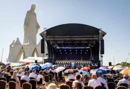Widok na scenę z chórem, z perspektywy widocznej na zdjęciu publiczności, w tle Pomniik Matki Boskiej