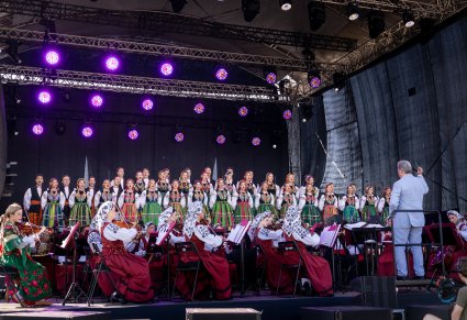 Orkiestra w kostiumach wilanowskich i podhalańskich, na czele z dyrygentem, Dyrektorem zespołu, w tle chór w kostiumach łowickich