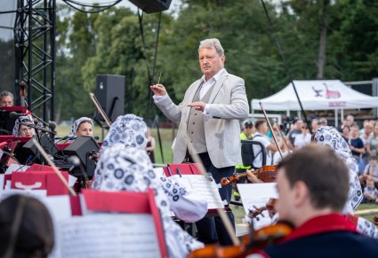Dyrektor zespołu, podczas dyrygowania na scenie, widoczna także część orkiestry w kostiumach wilanowskich oraz część publiczności w tle