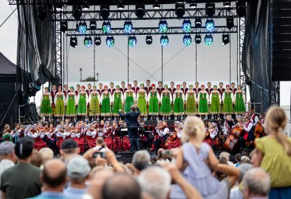 Widok na orkiestrę w kostiumach wilanowskich i podhalańsklich, razem z drygentem, a także na chór w kostiumach kurpiowskich, z perspektywy publiczności