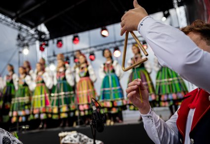 Zbliżenie na muzyka orkiesty grajacego na trójkącię, w tle chór w kostiumach łowickich