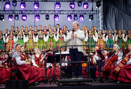 Dyrektor zespołu przemawiający podczas koncertu, za nim orkiestra w kostiumach wilanowskich oraz chór w kostiumach łowickich