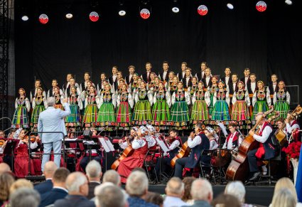 Na scenie chór w kostiumach łowickich, niżej orkiestra w kostiumach wilanowskich a przed nimi Dyrygent trzymający w górze batutę