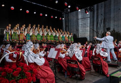 Na scenie Artyści Chóru w kostiumach łowickich, niżej Orkiestra w kostiumach wilanowskich a przed nią Dyrygent