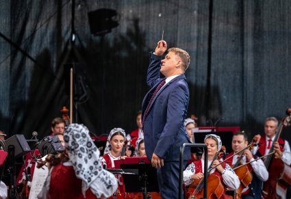 Zbliżenie na Dyrygenta, który w górze trzyma batutę, obok Artyści Orkiestry w kostiumach wilanowskich