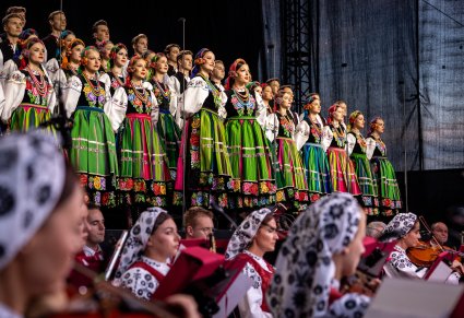 Na środku sceny dwie Solisty w kostiumach łowickich, za nimi reszta część chóru, niżej widoczna część orkiestry w kostiumach wilanowskich w trakcie grania na instrumentach