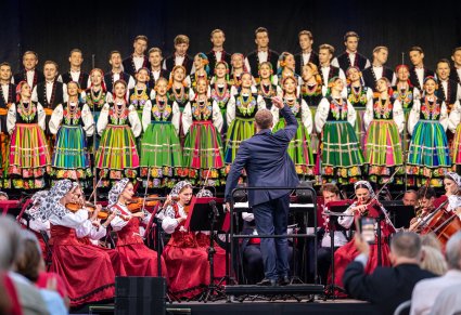 Na pierwszym planie Dyrygent zwrócony w stronę orkiestry w kostiumach wilanowskich i chóru w kostiumach łowickich