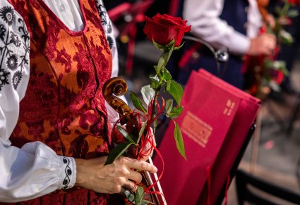 Zbliżenie na Artystę Orkiestry w kostiumie wilanowskim, trzymającą w dłoni czerwoną róże, w tle bordowa teczka na nuty ze złotym logotypem zespołu "Mazowsze"