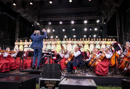 Widok na scenę plenerową na której znajduje się Chór w kostiumach stylowych z końca XIX w. wraz z Orkiestrą w kostiumach wilanowskich i Dyrygentem