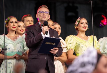 Zbliżenie na Konferansjera w garniturze, który trzyma w ręku podkładkę ze złotym logotypem "Mazowsze", za nim widoczna część Artystów Chóru w kostiumach stylowych z końca XIX w.