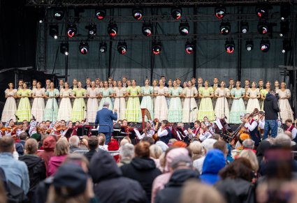 Na pierwszym planie na scenie Artyści Chóru w kostiumach stylowych z końca XIX w., niżej Artyści Orkiestry w kostiumach wilanowskich i Dyrygent trzymający batutę, widoczna część Publiczności
