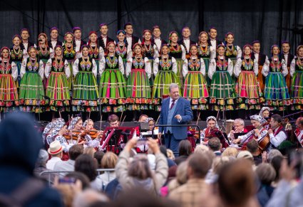 Na pierwszym planie widoczna część Publiczności skierowanej w stronę sceny na której znajduje się Dyrygent, Orkiestra w kostiumach wilanowskich i Chór w kostiumach łowickich