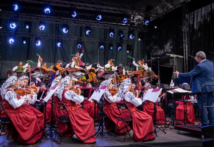 Na pierwszym planie Artyści Orkiestry w kostiumach wilanowskich wraz z Dyrygentem trzymającym w ręku batutę, wyżej na scenie Artyści w kostiumach ludowych podczas finałowej choreografii