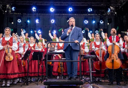 Na pierwszym planie Dyrektor zespołu wypowiadający się do mikrofonu, w tle widoczni Artyści orkiestry w kostiumach wilanowskich, a wyżej na scenie Artyści Chóru w kostiumach sannickich