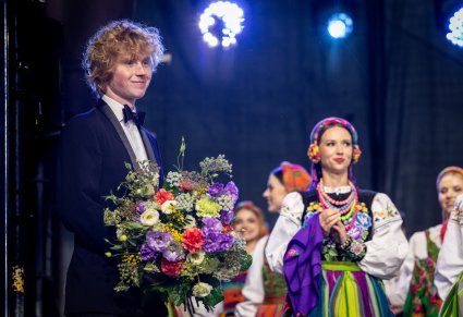 Zbliżenie na młodego Pianistę, który trzyma w ręku otrzymane kwiaty, w drugim planie widoczni Artyści Chóru w kostiumach łowickich