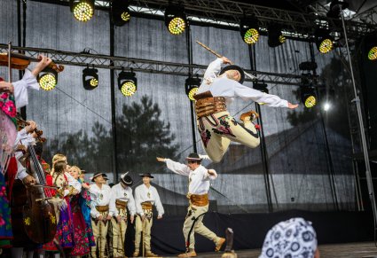 Na pierwszym planie Artysta w kostiumie podhalańskich wykonujący imponujący skok wraz z ciupagą, za nim część Artystów w kostiumach podhalańskich grająca na instrumentach