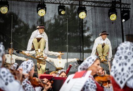 Dwóch Artystów w kostiumach podhalańskich w trakcie wykonywania impozujacej choreografii tanecznej w stylu góralskim, widoczna część Orkiestry w kostiumach wilanowskich