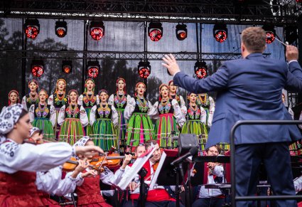Na pierwszym planie Dyrygent zwrócony w stronę Orkiestry i Chóru, Orkiestra jest w kostiumach wilanowskich, a Chór w kostiumach łowickich