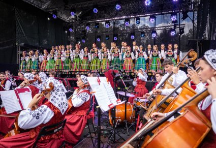 Na pierwszym planie część Orkiestry w kostiumach wilanowskich grająca na instrumentach, wyżej na scenie Artyści Chóru w kostiumach łowickich