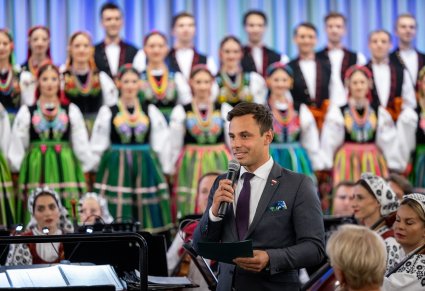 Przemawiający przedstawiciel samorządu, w tle chór w kostiumach łowickich