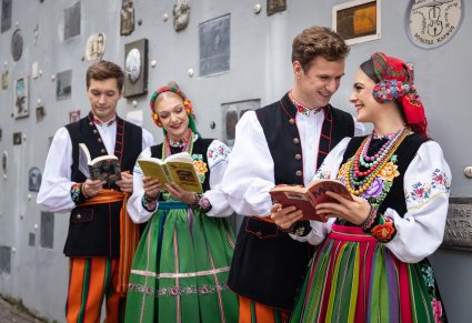 Dwie pary Artystów w kostiumach łowickich z książkami na tle charakterystycznej ściany na ul. Literackiej