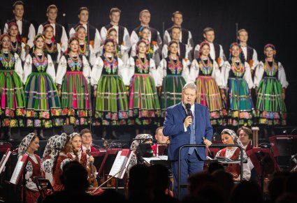 Dyrektor zespołu w trakcie przemowy do mikrofonu, za nim Orkiestra w kostiumach wilanowskich i Artyści chóru w kostiumach łowickich