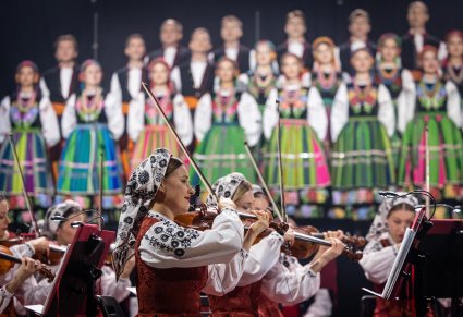 Na pierwszym planie Artystki Orkiestry w kostiumach wilanowskich, grające na instrumentach smyczkowych, w drugim planie Artyści Chóru w kostiumach łowickich