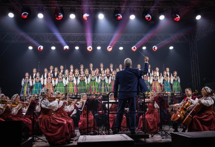 Na pierwszym planie Dyrygent zwrócony w stronę Orkiestry i Chóru, trzymający w górze batutę, Orkiestra w kostiumach wilanowskich, chór w kostiumach łowickich