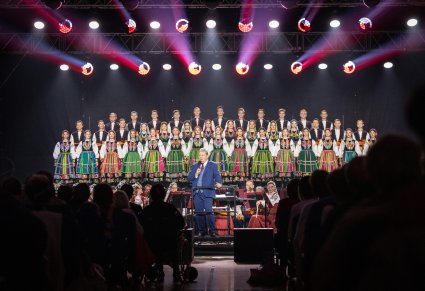 Na pierwszym planie Dyrektor zespołu przemawiający do mikrofonu, w tle siedząca orkiestra w kostiumach wilanowskich i Artyści Chóru w kostiumach łowickich