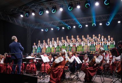 Na scenie Artyści Chóru w kostiumach łowickich, niżej Artyści Orkiestry w kostiumach wilanowskich oraz Dyrygent- Dyrektor zespołu "Mazowsze"