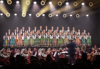 Na scenie Artyści Chóru w kostiumach łowickich w trakcie śpiewu, niżej sceny Artyści Orkiestry w kostiumach wilanowskich w trakcie grania na instrumentach oraz Dyrygent zwrócony w stronę Orkiestry i Chóru, trzymający w ręku batutę
