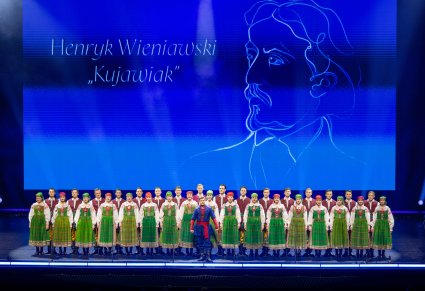Na pierwszym planie Solista w kostiumie kujawskim, za nim na drugim planie stojący w dwóch rzędach Artyści Chóru w kostiumach kurpiowskich