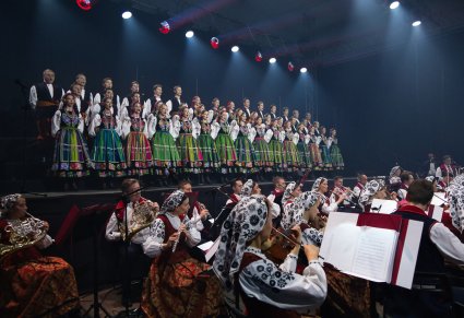 widok na scenę na której znajdują się Artyści Chóru w kostiumach łowickich, poniżej Artyści Orkiestry w kostiumach wilanowskich