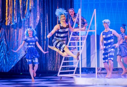 Na pierwszym planie postać trójkąta w niebieskim kostiumie, skacząca przez dużą skakankę, w tle reszta część postaci trójkątów a na drabinie jedna z postaci Gwiazd