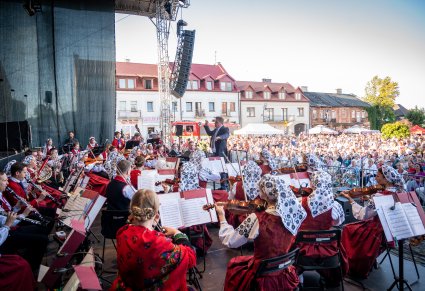 Orkiestra widoczna z boku wraz z Dyrygentem a w tle widać publiczność i budynki