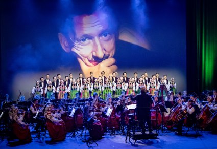 Dyrektor Jacek Boniecki zwrócony do Orkiestry, za Orkiestrą chór ustawiony w rzędy w kostiumach łowickich na ekranie postać Tadeusza Sygietyńskiego