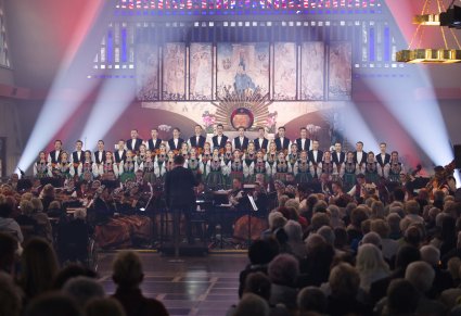 Przed podświetlonym ołtarzem Artyści Chóru w kostiumach łowickich, widoczni również Artyści Orkiestry w kostiumach wilanowskich oraz Dyrygent