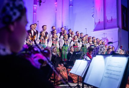 Na pierwszym planie Muzyczka w kostiumie wilanowskim grająca na instrumencie smyczkowym, w tle śpiewający Chór w kostiumach łowickich