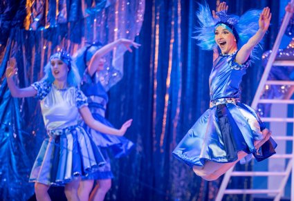 Na pierwszym planie skaczące postaci trójkątów w niebieskich kostiumach i niebieskich perukach