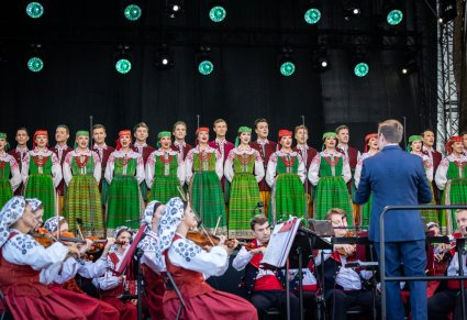 W tle chór w kostiumach kurpiowskich, z przodu dyrygent zespołu, poniżej część orkiestry w kostiumach wilanowskich.