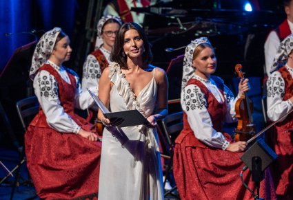 Zbliżenie na konferansjerkę w długiej, białej sukni, za nią Muzycy z orkiestry