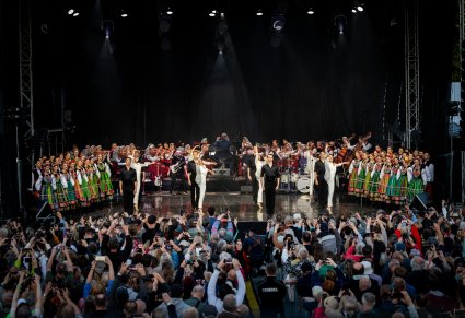 Widok na całą scenę i Publiczność. Na scenie Artyści Baletu w kostiumach współczesnych wykonujący choreografię taneczną, po bokach sceny widoczni Artyści chóru w kostiumach łowickich a w tle Orkiestra w kostiumach wilanowskich wraz z Dyrygentem