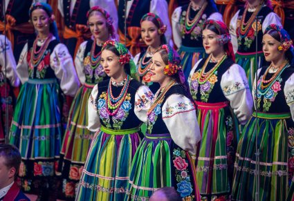 Na pierwszym planie dwie Solistki w trakcie śpiewu, w tle część Artystów Chóru w kostiumach łowickich