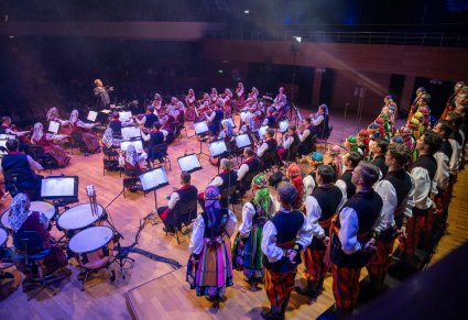 Na scenie Artyści Chóru w kostiumach łowickich ustawieni w 3 rzędy, niżej Orkiestra w kostiumach wilanowskich z Dyrektorem zespołu "Mazowsze", który dyryguje koncert
