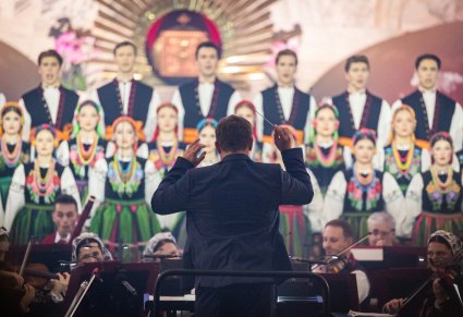 Zbliżenie na sylwetkę Dyrygenta z batutą w ręku, przed nim Artyści Orkiestry w kostiumach wilanowskich oraz Artyści Chóru w kostiumach łowickich
