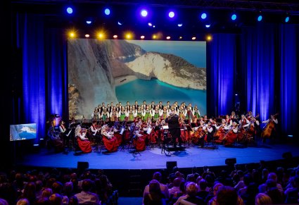 Widoczne pierwsze rzędy publiczności, na scenie Dyrektor Jacek Boniecki dyrygujący koncert, Orkiestra a za nią chór w rzędach w kostiumach łowickich