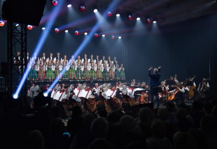 Na podświetlonej scenie znajdują się Artyści Chóru w kostiumach łowickich, Artyści Orkiestry w kostiumach wilanowskich oraz Dyrygent trzymający w górze batutę