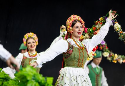 Zbliżenie na grupę Artystów w kostiumach lubuskich podczas wykonywania choreografii tanecznej wraz z rekwizytami, w tym przypadku są to kwiaty