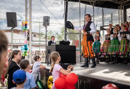 Na scenie na pierwszym planie Solista w trakcie wykonywania utworu "Furman" w tle część Artystów Chóru w kostiumach łowickich, widoczny również Akompaniator grający na instrumencie, przy scenie Publiczność