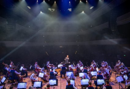 Na pierwszym planie Dyrektor zespołu "Mazowsze" trzymający w ręku batutę i zwrócony w stronę Orkiestry w kostiumach wilanowskich